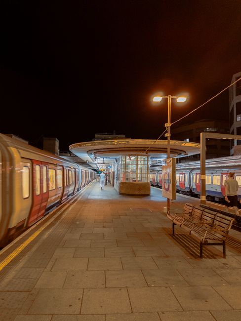 A nighttime scene at a train station platform shows a pair of train carriages stationary on either side of the platform, with one carriage painted in red and cream and the other in blue with red and w