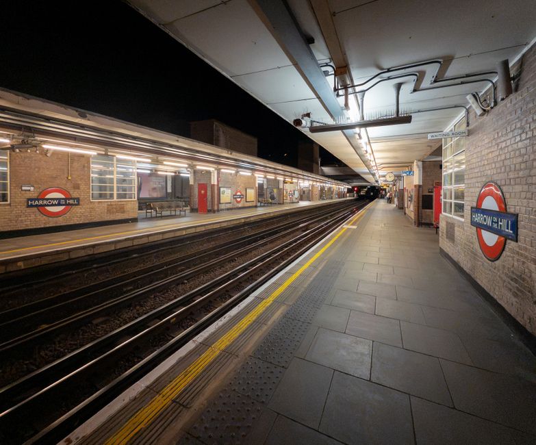 A nighttime scene at a train station platform shows a pair of train carriages stationary on either side of the platform, with one carriage painted in red and cream and the other in blue with red and w