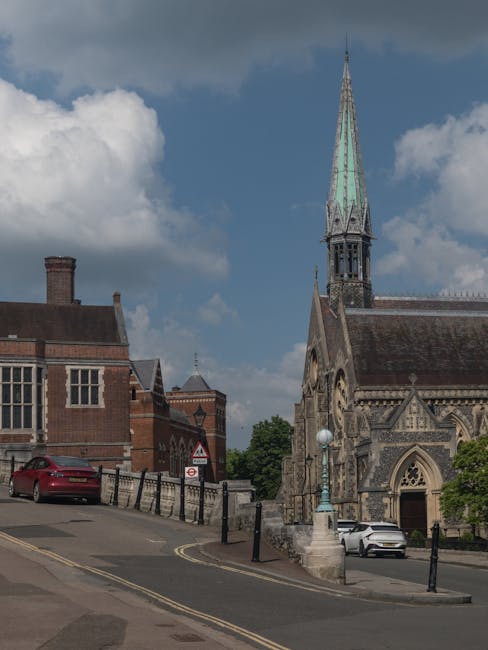 A narrow street in North Harrow with a church featuring a tall, pointed spire and intricate stone architecture visible on the right side. The street has a slight uphill gradient with a black metal rai