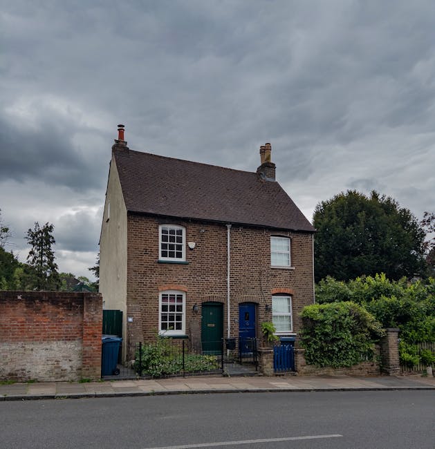 A two-storey brick house situated on a residential street, with a dark tiled roof, two visible chimneys, and white-framed windows. The front façade features two doors, one green and one blue, each ac
