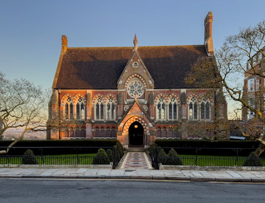 A two-storey brick house situated on a residential street, with a dark tiled roof, two visible chimneys, and white-framed windows. The front façade features two doors, one green and one blue, each ac